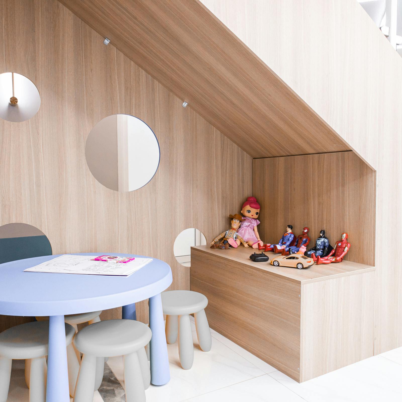 A bright, lived-in children's play corner with a soft blue play table, child-sized stools, and a built-in wooden toy niche